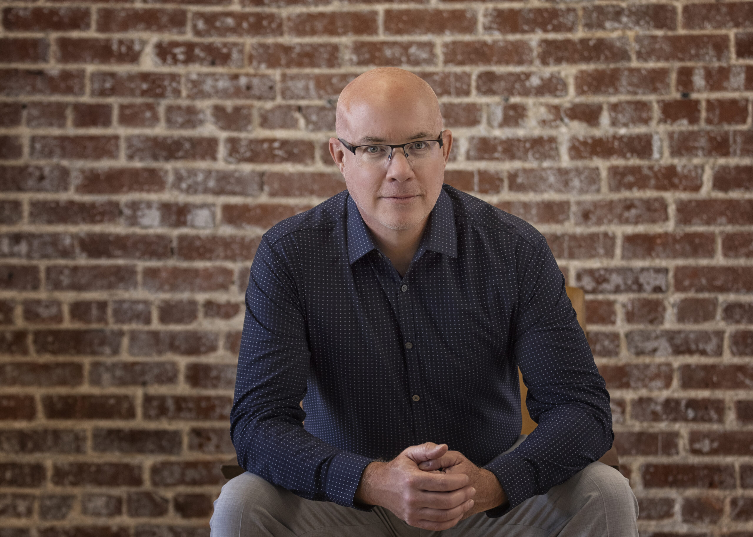 Portrait of Chis Duncan leaning forward in a chair hands clasped out front looking fearless in a black button down and redbrick wall behind him.
